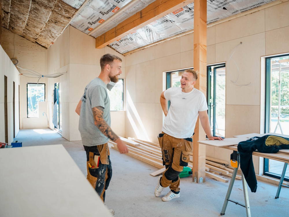 To tømrere står og snakker smilende inde i et hus, hvor væggene er under isolering.